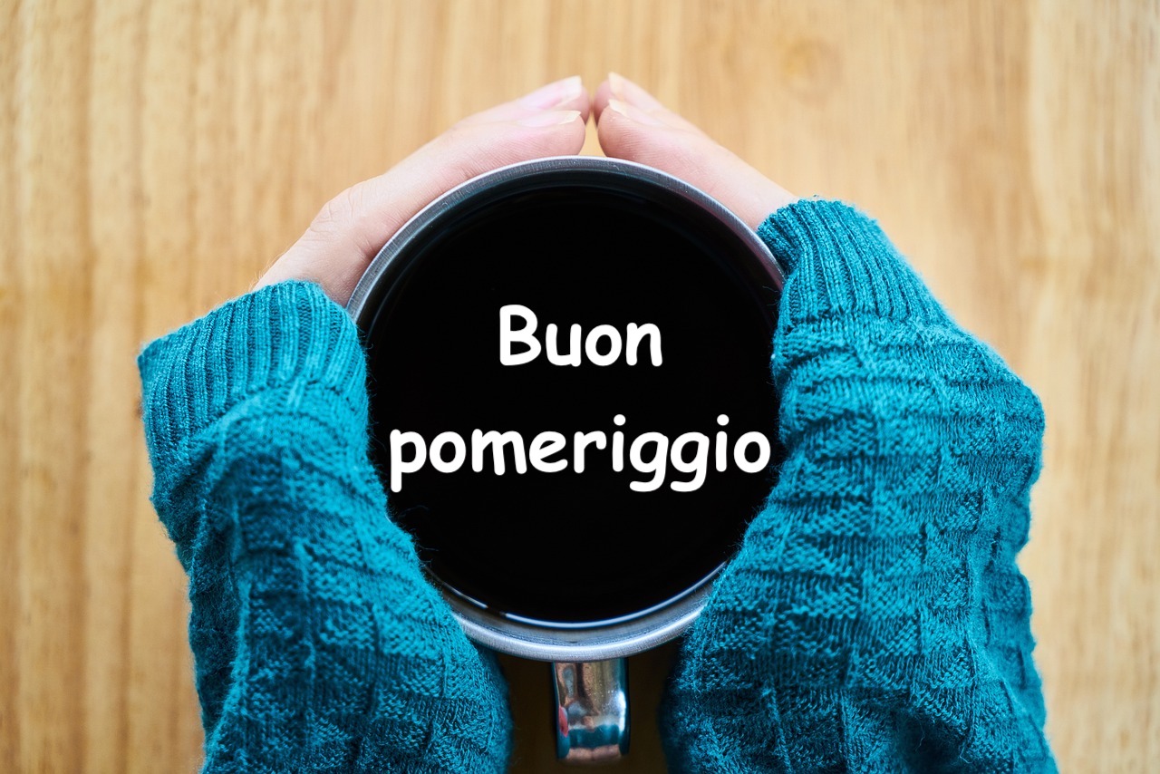 le mani di una donna con un maglione invernale stringono una tazza di caffè con la scritta buon pomeriggio all'interno della tazzina  